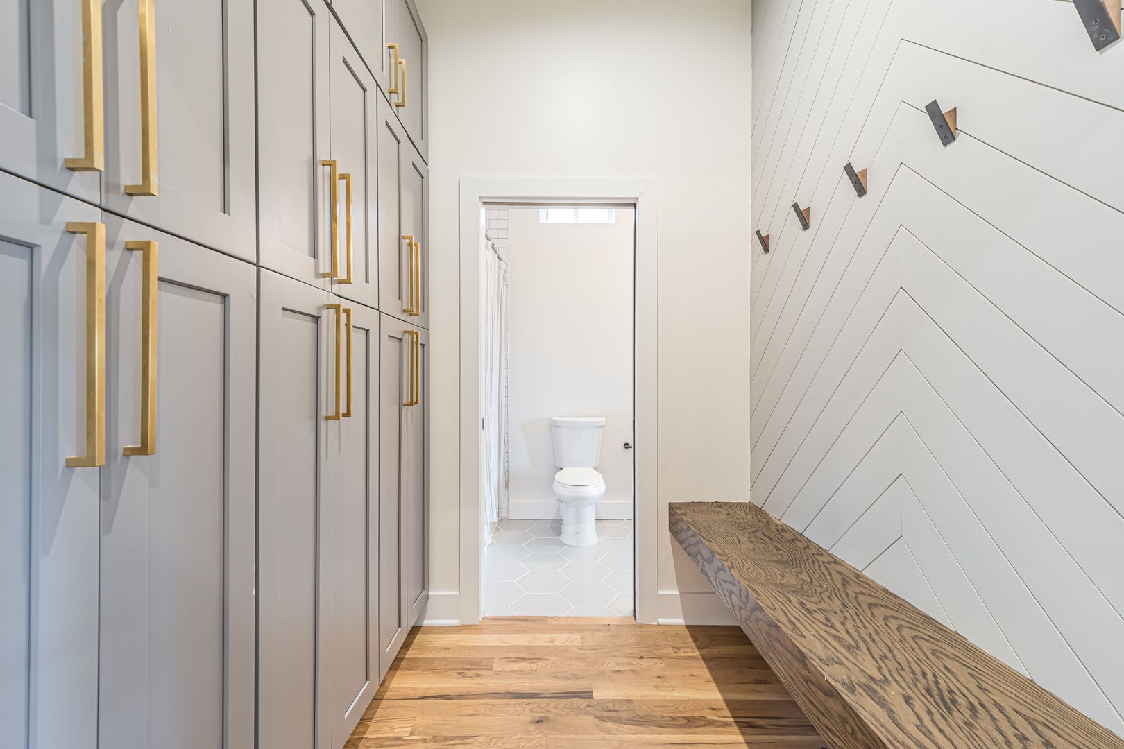 Narrow modern hallway with gray cabinets, wood floor, white wall benches, and a doorway to a bathroom.