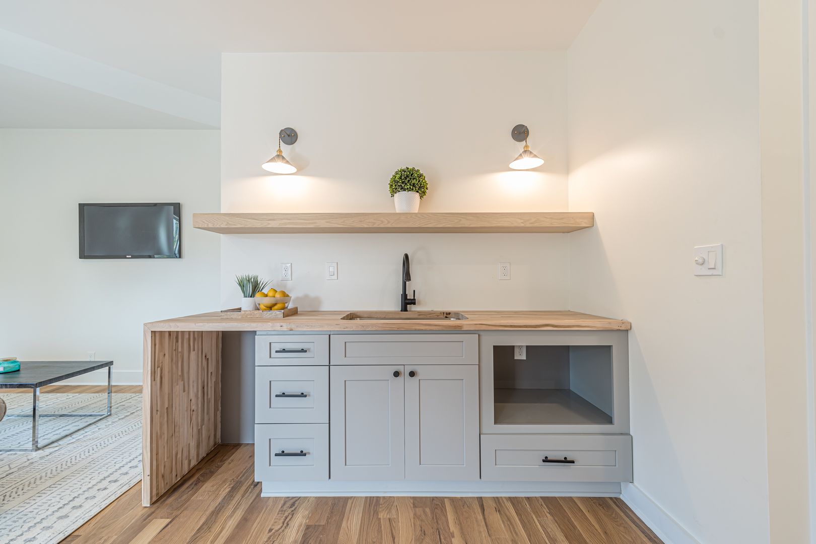 Modern white kitchen nook with wood countertops, sink, open shelf, and small potted plant under two wall lights