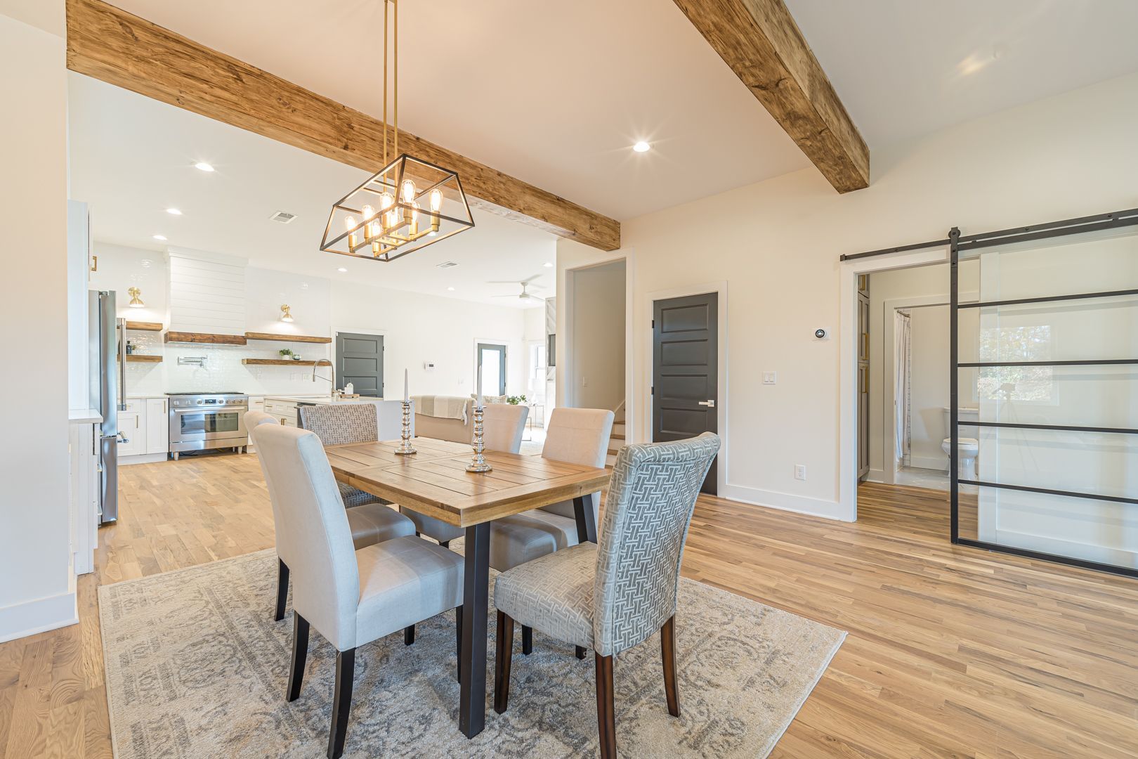 Bright open dining room with wood beams, long table, upholstered chairs, and kitchen visible in the background