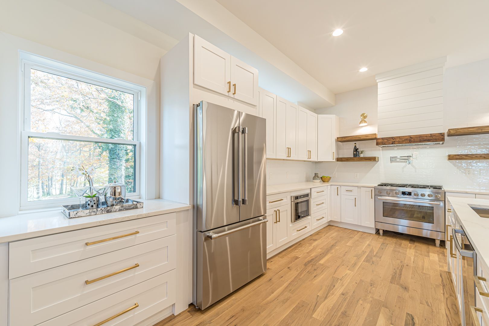 Bright white kitchen with stainless steel appliances, wood floors, and a large window over the sink