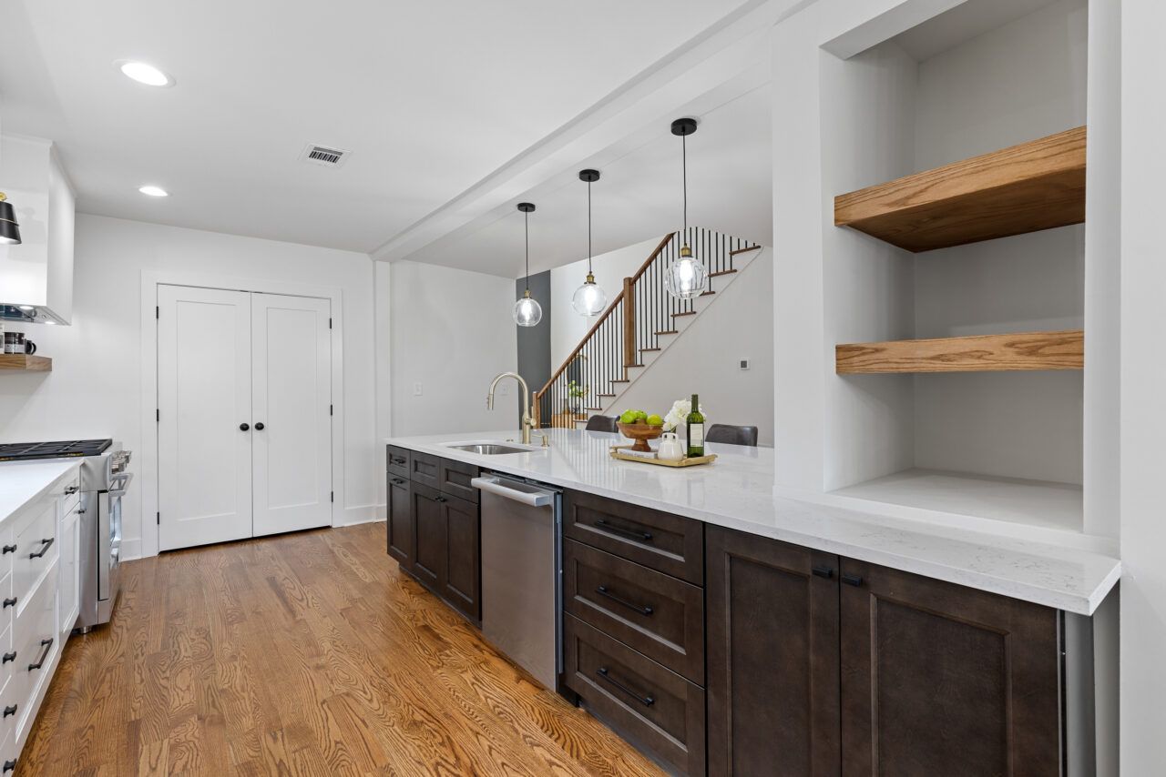 Modern kitchen with dark cabinets, white countertops, hardwood floors, and open shelves near a staircase