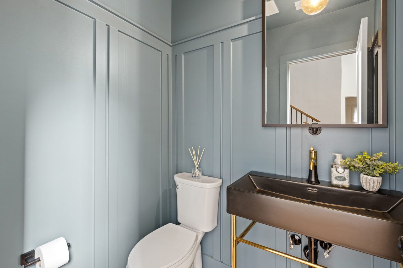 Small bathroom with pale blue walls, toilet, vanity sink, mirror, and gold accents