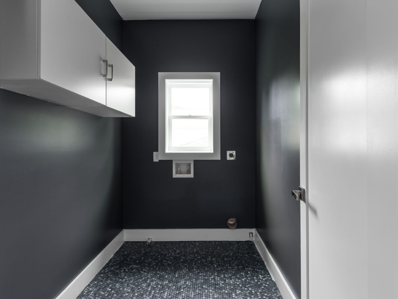 Small gray laundry room with a window, cabinets, and speckled floor.