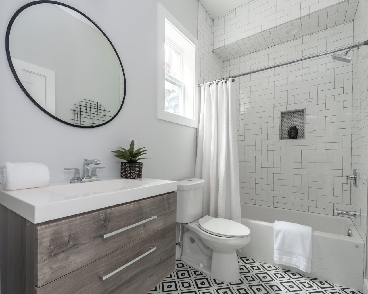 Bright white bathroom with round mirror, wood vanity, toilet, tub, and patterned black-and-white floor tiles.