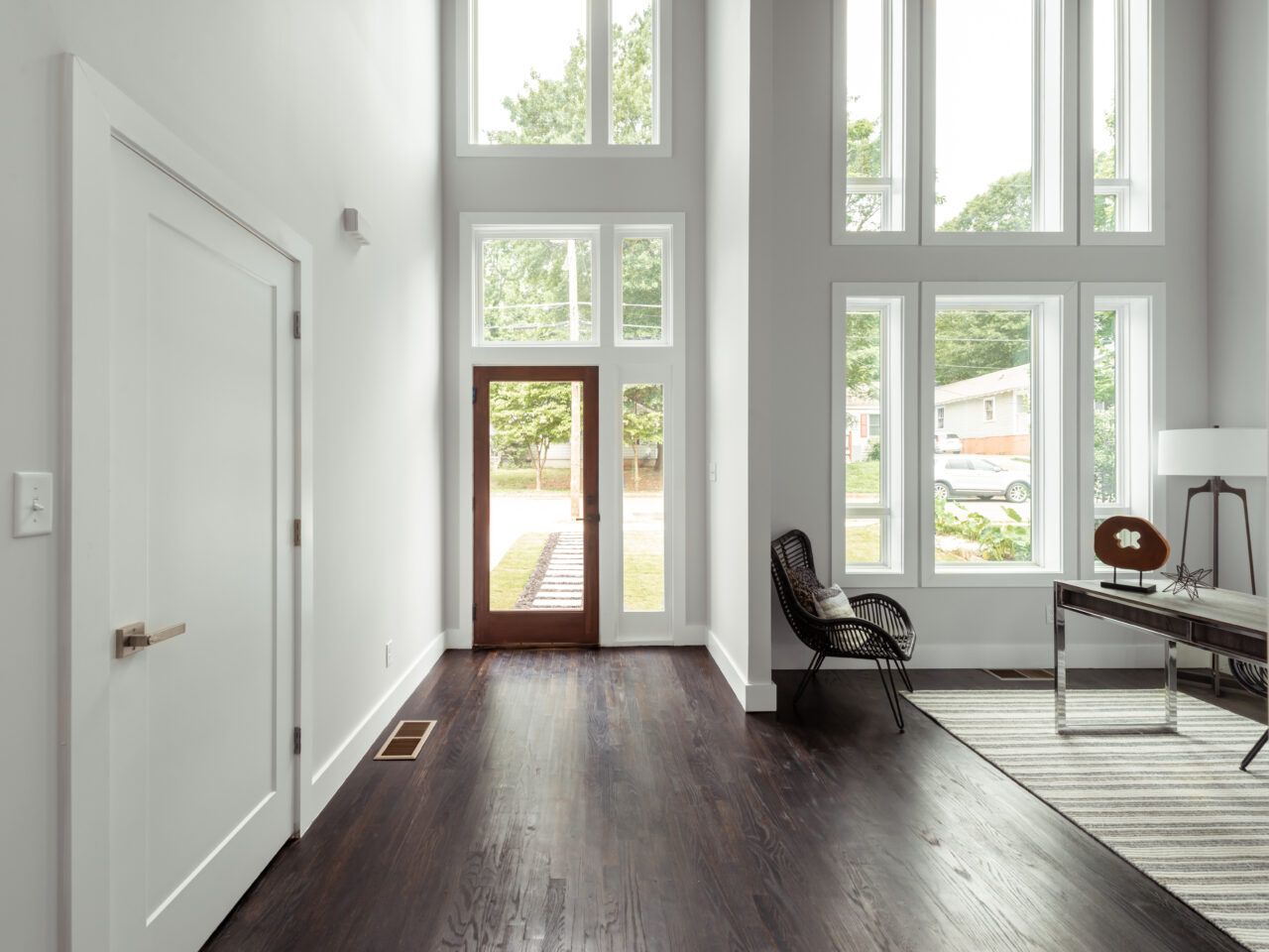 Bright modern foyer with tall windows, dark wood floors, a white door, and a chair near the entry.