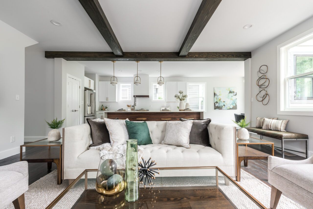 Bright modern living room with white sofa, glass coffee table, exposed beams, and open kitchen beyond