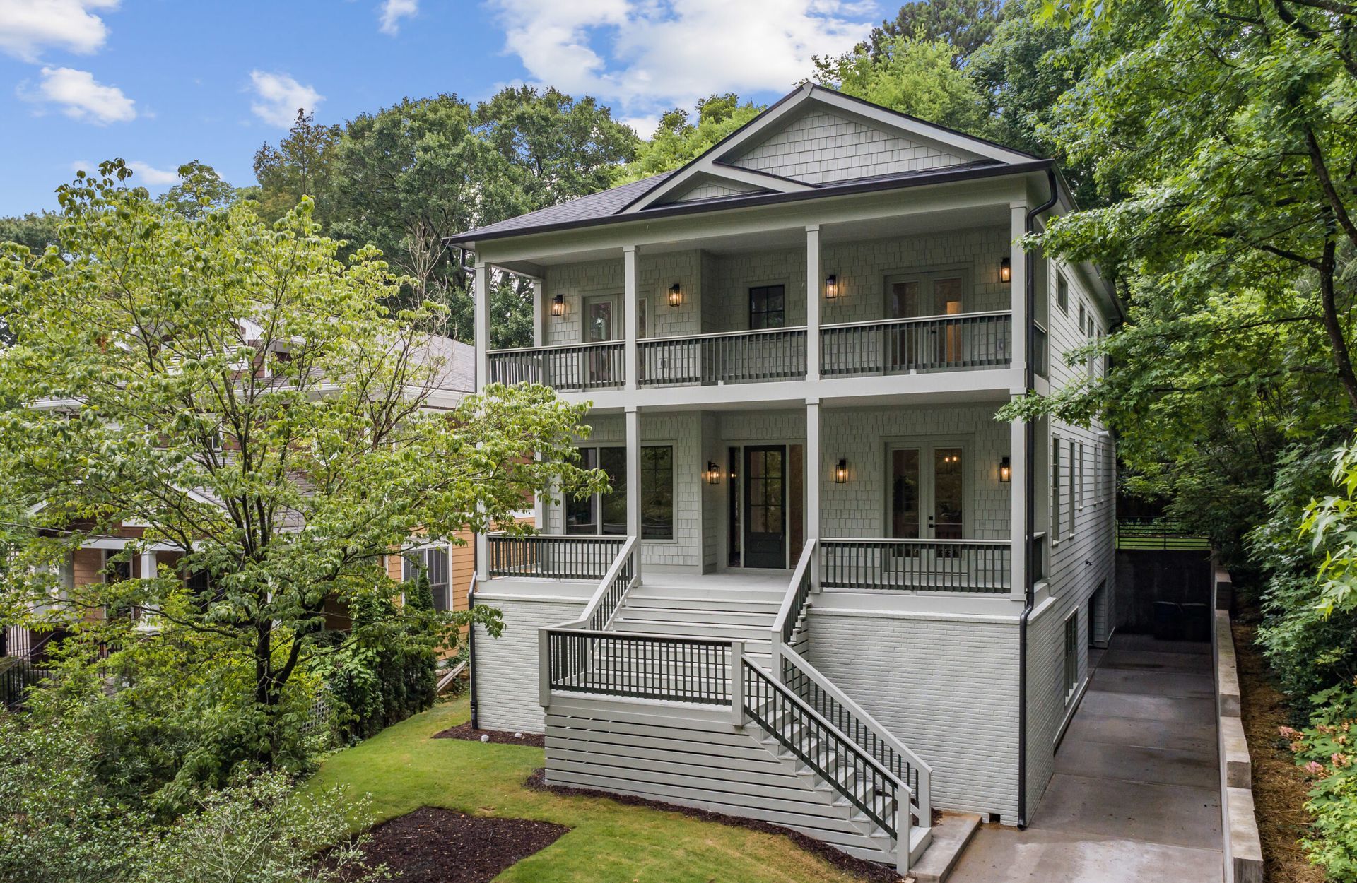 White two-story house with wraparound porch and stairs, surrounded by trees and greenery