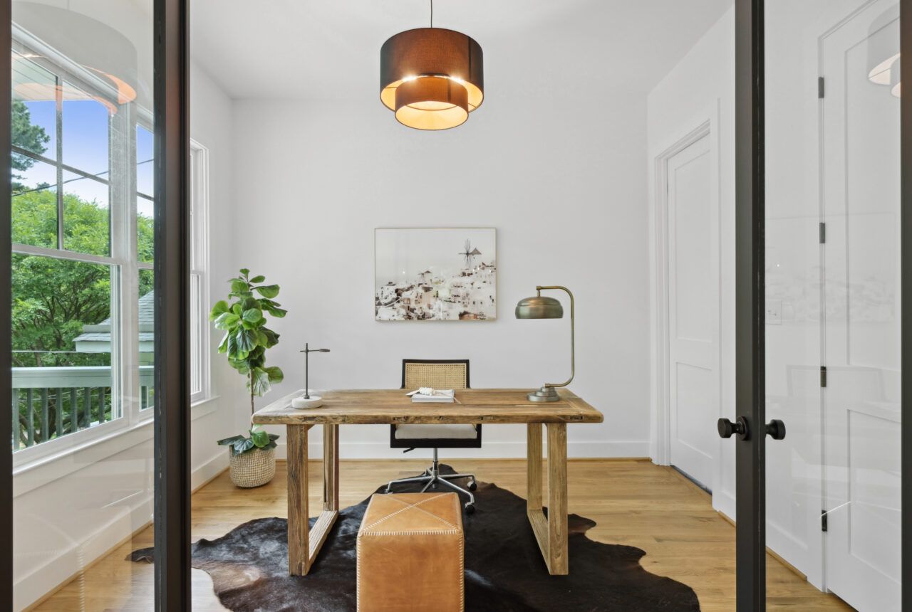 Bright home office with wooden desk, chair, plant, lamp, and large window with a porch view.