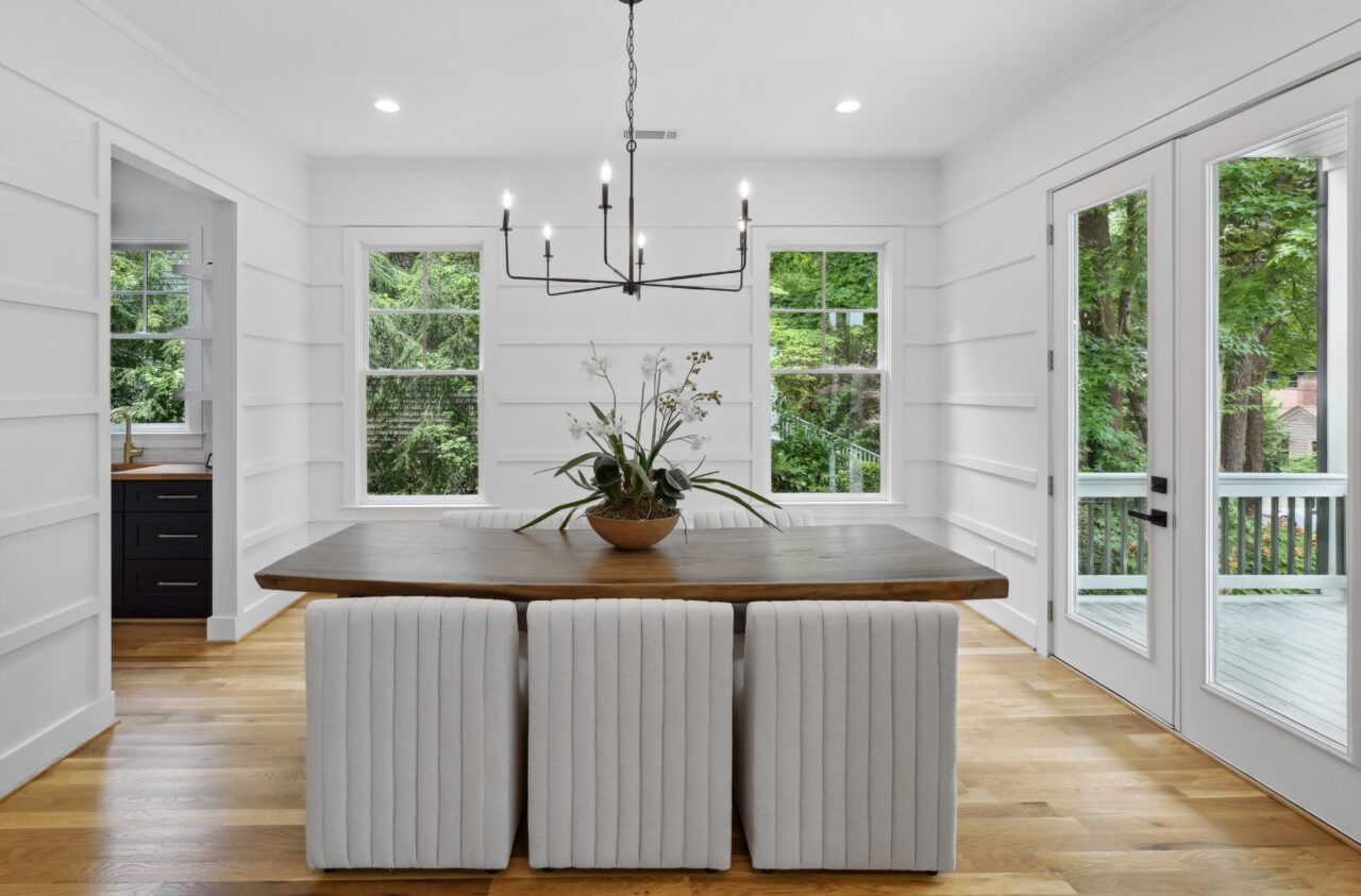 Bright dining room with a wood table, white chairs, black chandelier, and large windows overlooking greenery