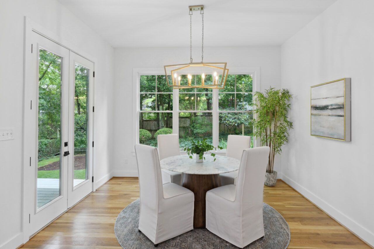 Bright dining room with white chairs, round table, chandelier, and large windows overlooking greenery