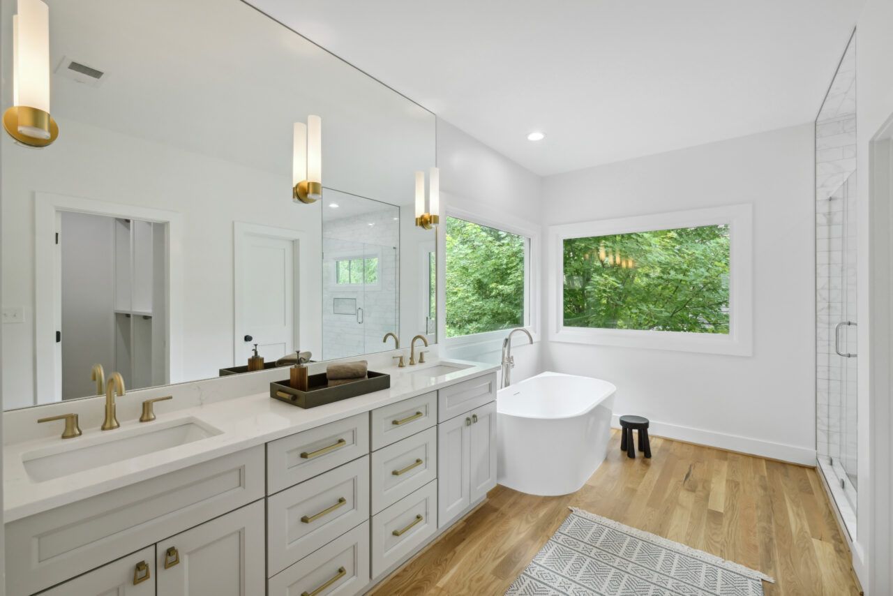 Bright modern bathroom with white vanity, freestanding tub, wood floor, and large windows showing greenery