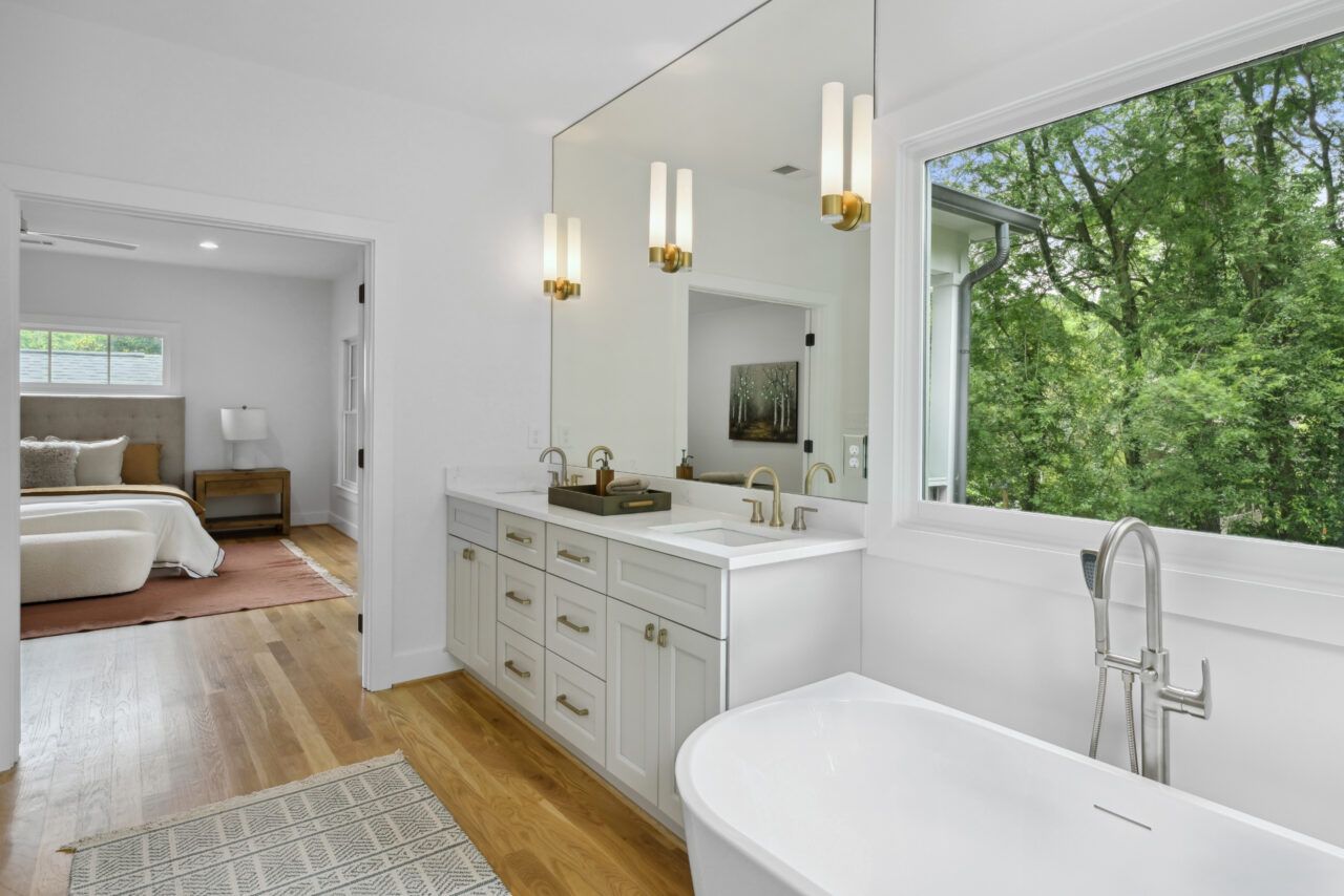 Bright bathroom with white vanity, freestanding tub, and large window overlooking green trees