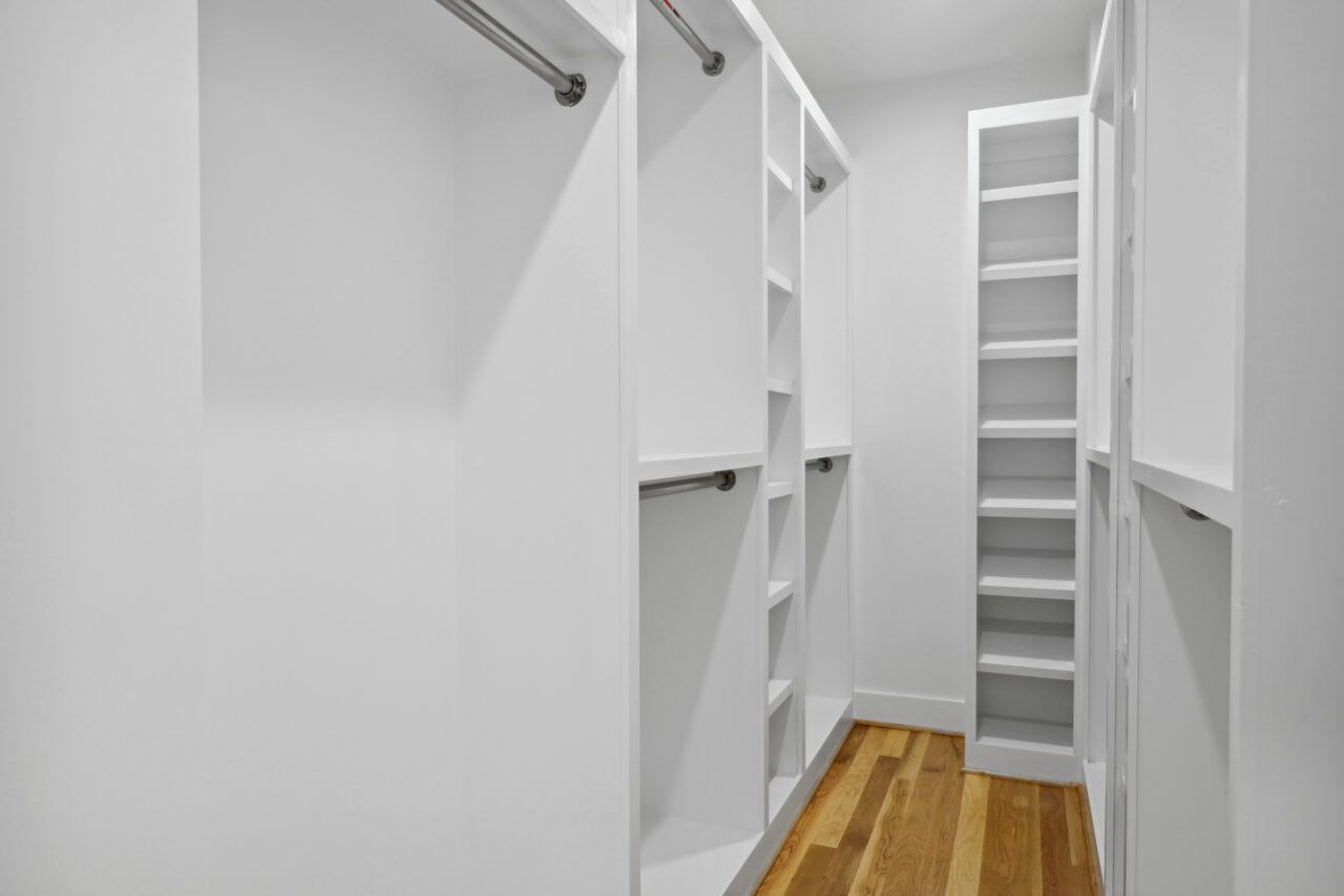 Narrow white walk-in closet with built-in shelves, hanging rods, and wood floor.