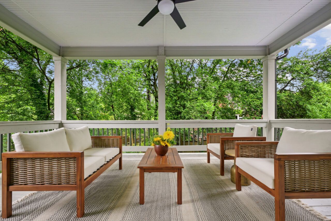 Covered porch with wicker seating, a coffee table, and green wooded view beyond the railing