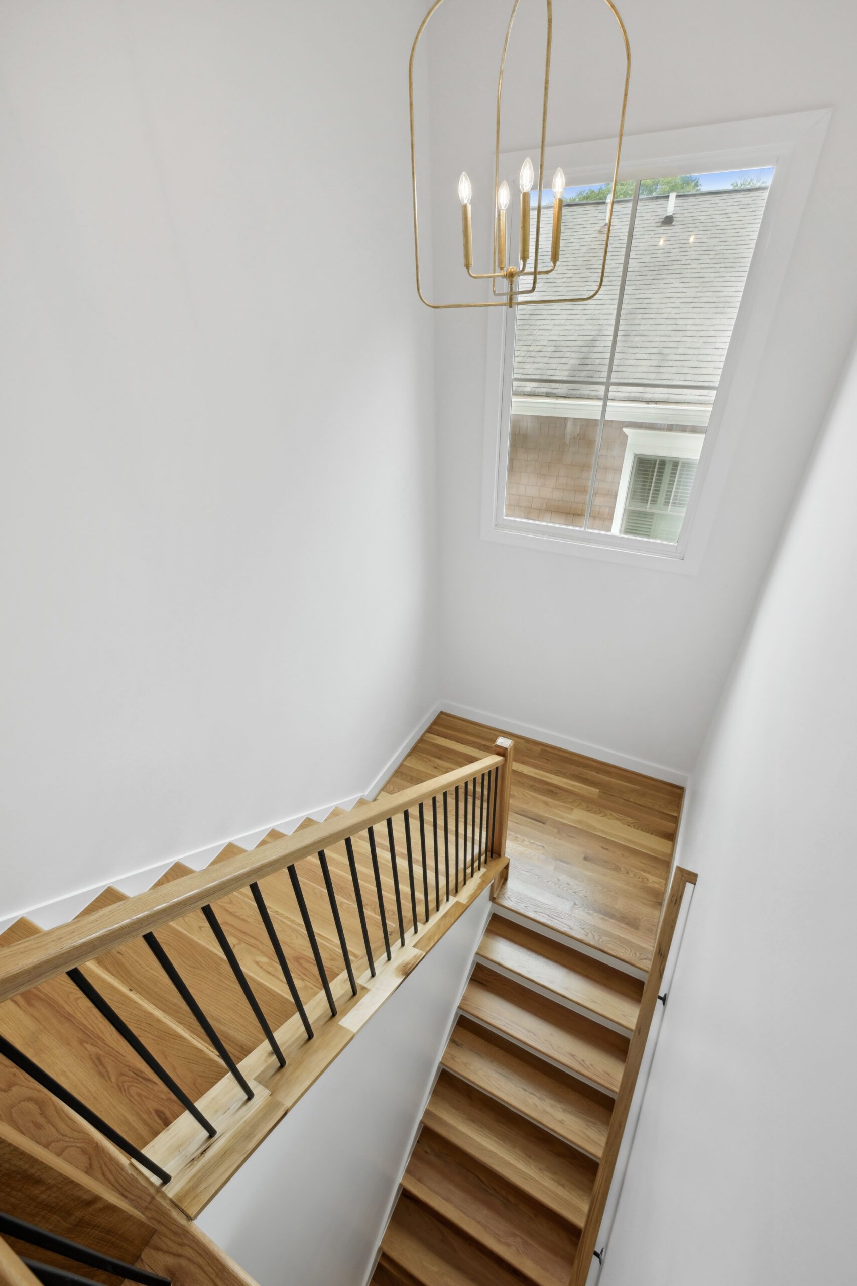 Staircase with wooden steps and railing in a white interior, lit by a modern chandelier near a window.