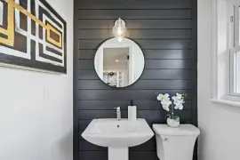 Modern bathroom with white pedestal sink, round mirror, black shiplap wall, and floral decor