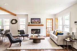 Bright living room with fireplace, gray sofa, black chairs, coffee table, and large windows
