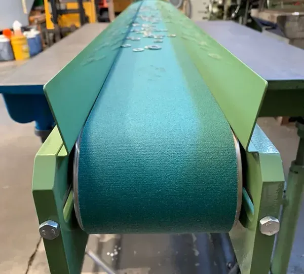 Vista ravvicinata di un nastro trasportatore industriale verde con sponde laterali in metallo, all'interno di un'officina.
