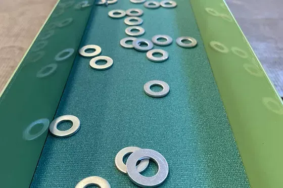 Primo piano di rondelle metalliche sparse su un nastro trasportatore industriale verde.