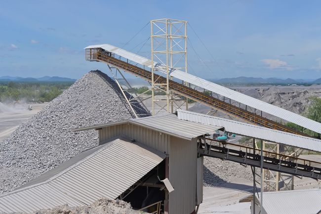 In una cava industriale, sotto un cielo azzurro e limpido, un sistema di nastri trasportatori convoglia roccia frantumata verso un grande cumulo.