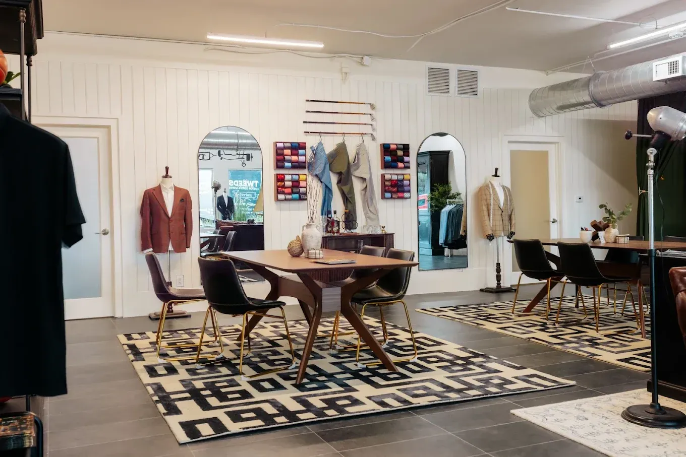 Interior of a clothing store with suits and tables. White walls, patterned rug, and arched mirrors.