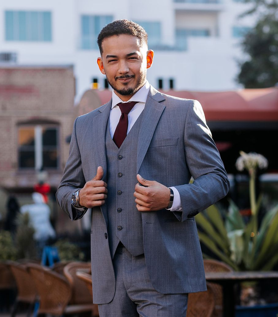 Man in red suit, blue patterned shirt, posing outdoors near a building with a car and greenery.