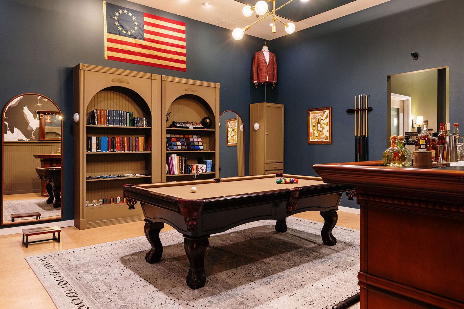 Game room with pool table, bookshelves, bar, and American flag. Dark blue walls and a rug.