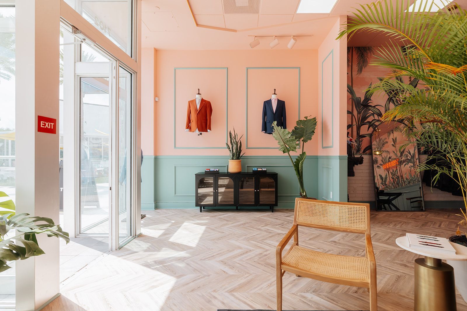A brightly lit clothing store interior with mannequins displaying suits, pink and teal walls, and a rattan chair.