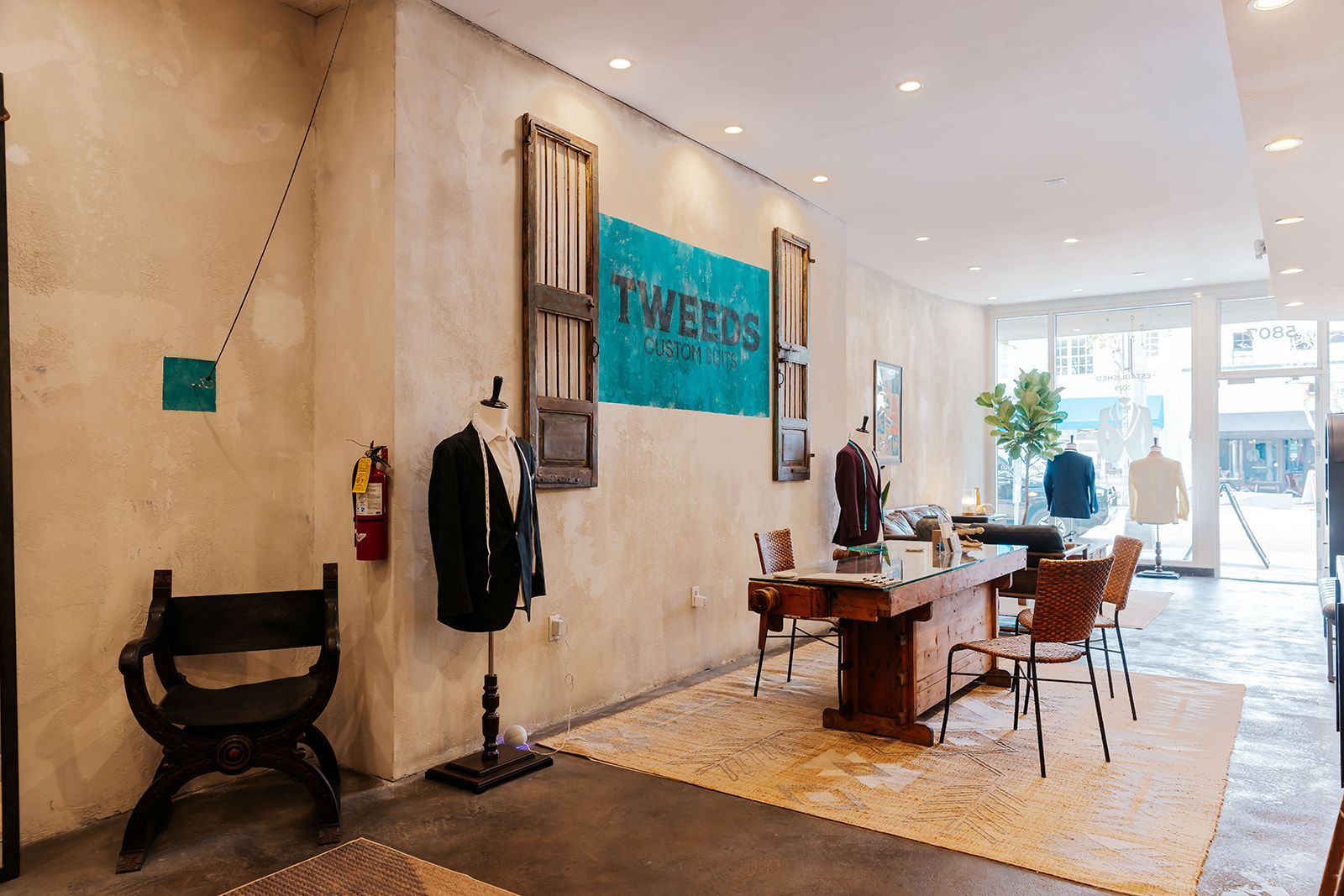 Interior of a menswear shop. Suits on display, a large wooden table with chairs, and a turquoise sign on a textured wall.