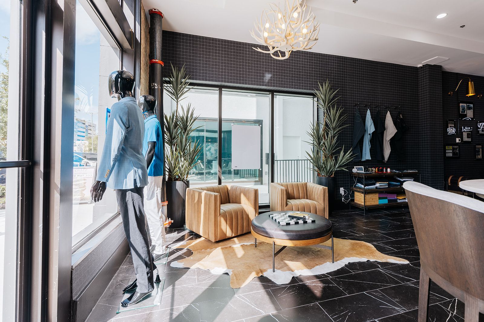 Men's clothing store interior with mannequins, seating area, and large windows. Dark walls, gold accents.