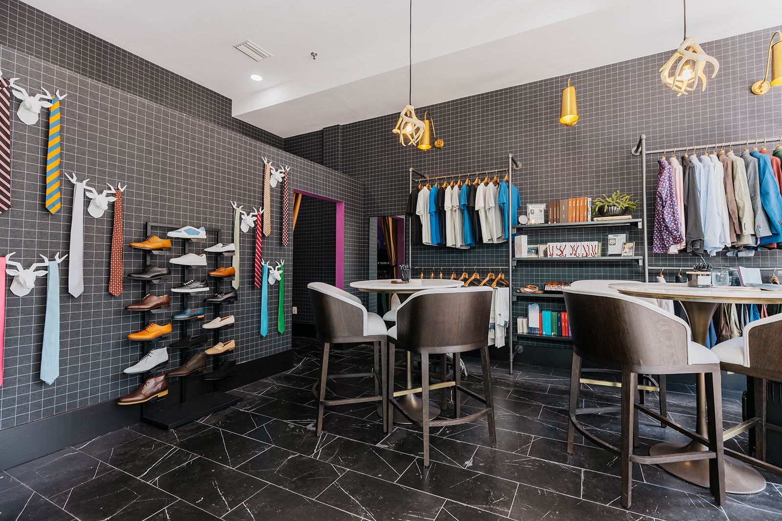 Men's clothing store interior with ties, shoes, and clothing racks. Black tile floor, neutral chairs.