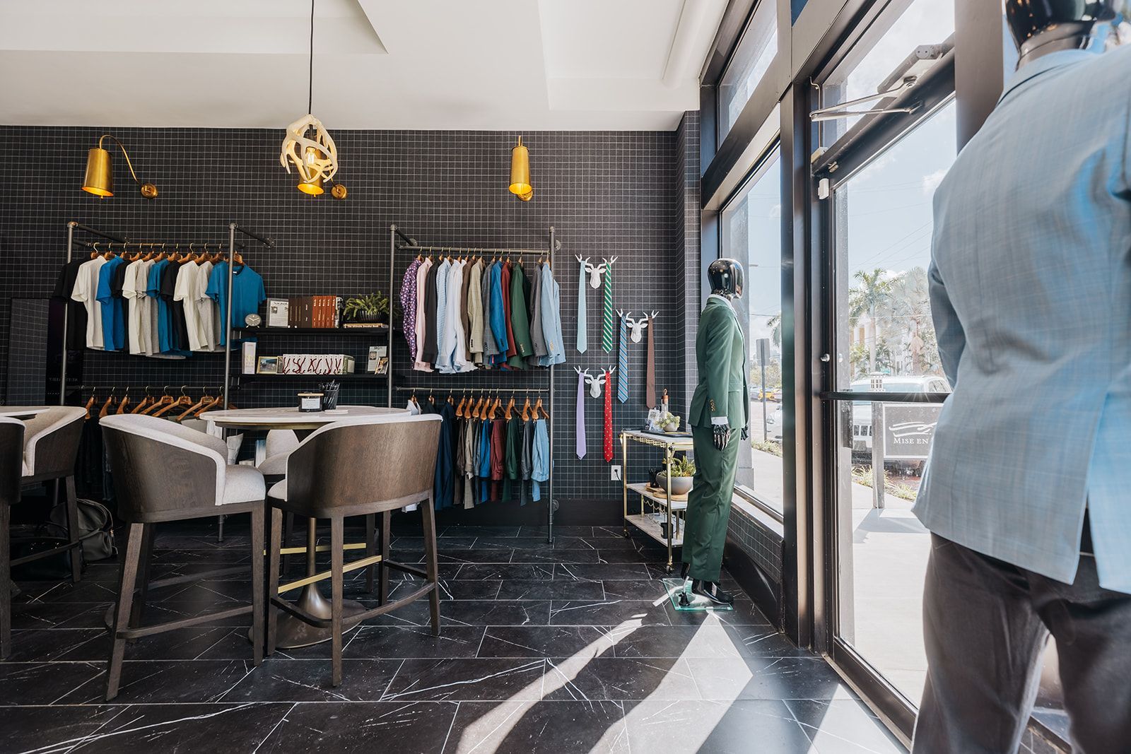 Interior of a clothing store with racks of shirts and suits, display mannequins, and a table with chairs.