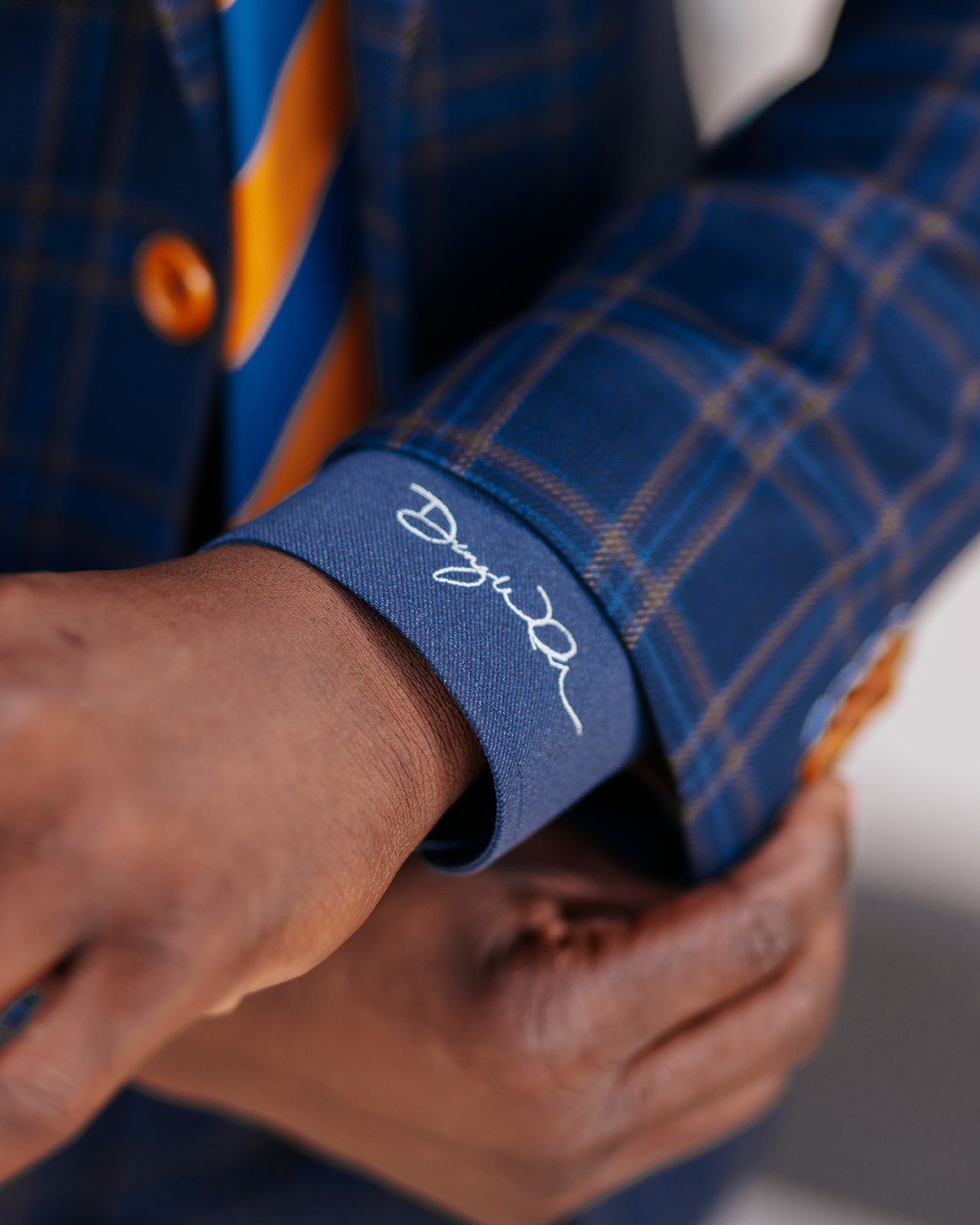 Close-up of a person adjusting a blue plaid suit sleeve, showing embroidered signature.