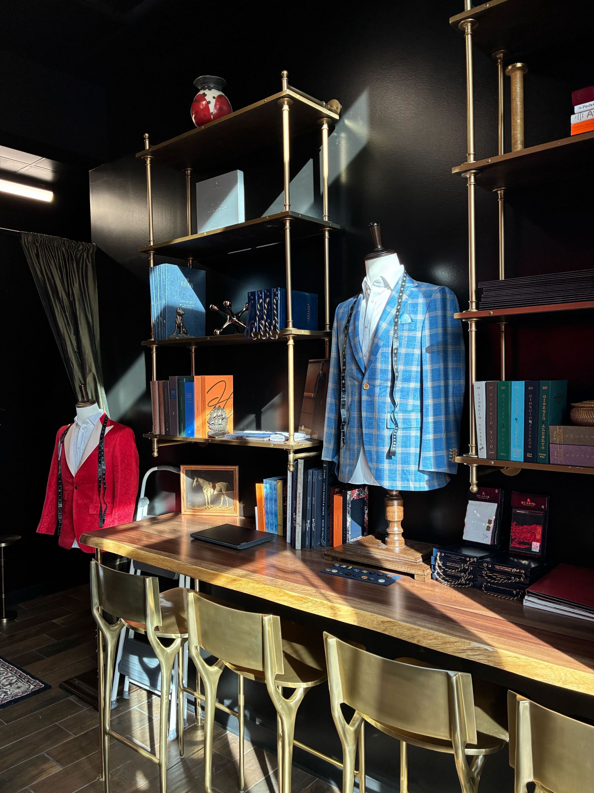 A boutique office with a wooden desk, gold chairs, and shelves holding suits, books, and accessories.