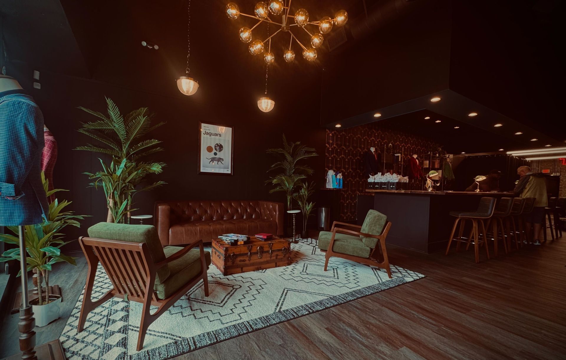 Lounge area with couch, chairs, plants, rug, and bar in a dimly lit space with a chandelier.