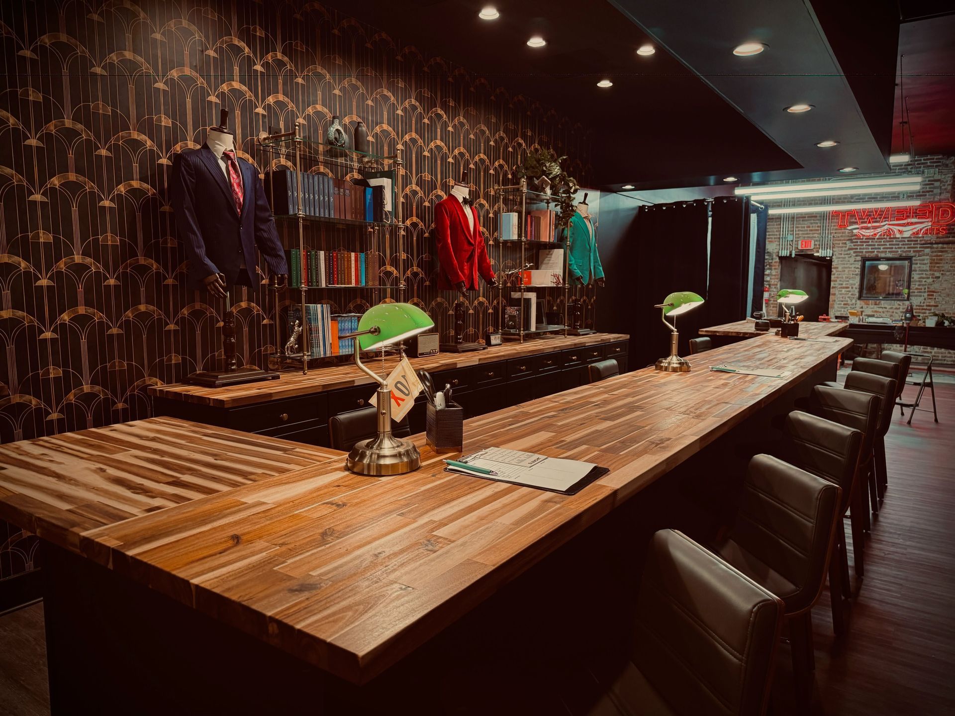 A dimly lit bar with a wooden counter, green lamps, and suits hanging on the patterned wall.