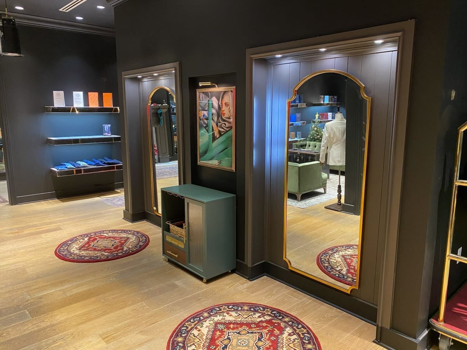 Interior of a clothing store with mirrors, colorful rugs, and a painting on a dark wall.