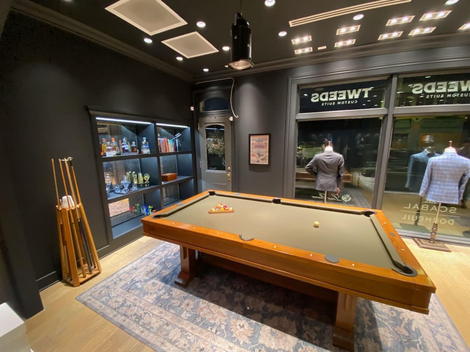 Pool table in a dark-walled shop, with suits visible outside. Cue rack and display case on the left.