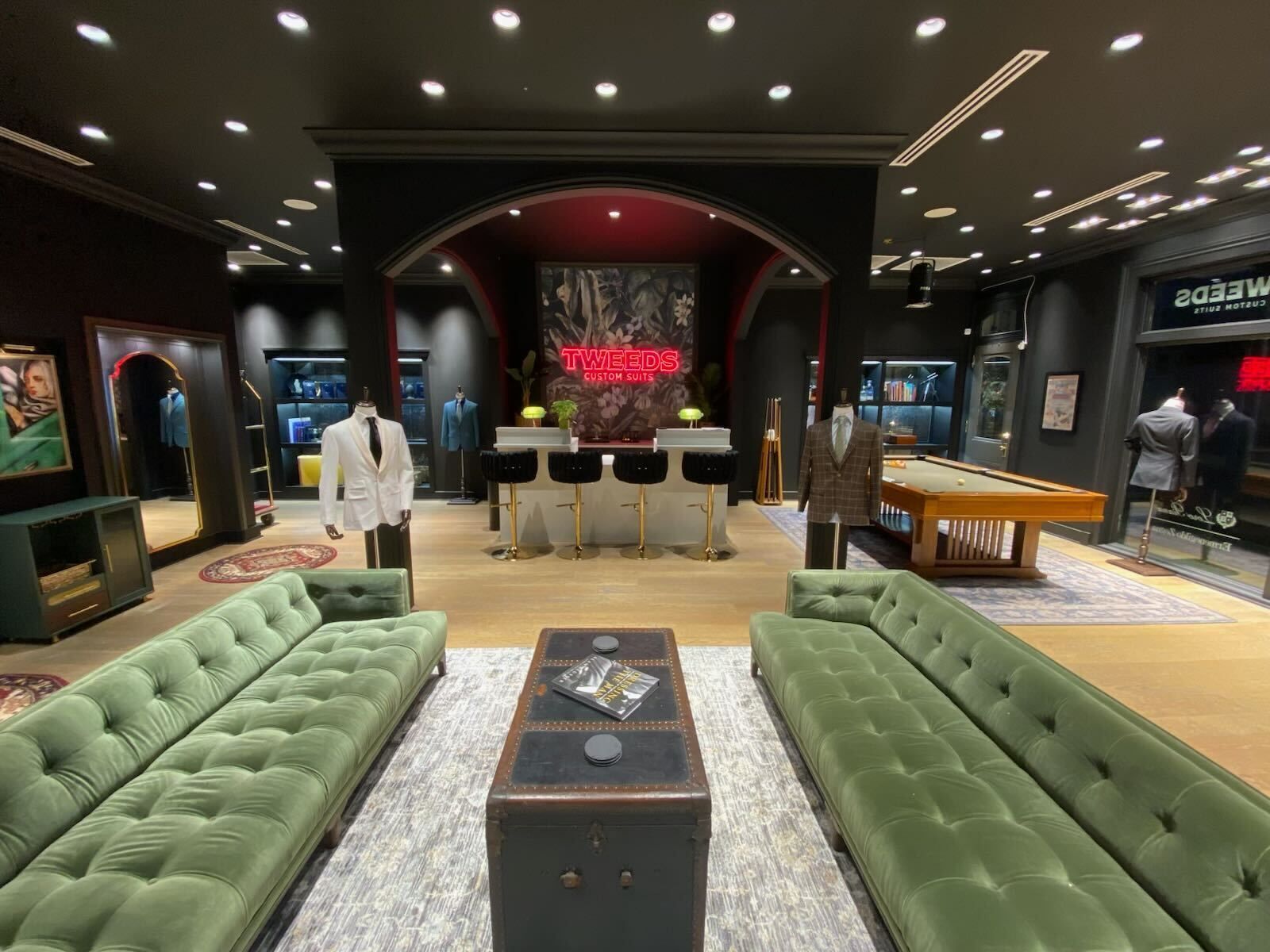 Upscale men's clothing store interior with green velvet sofas, suits on display, and a pool table.