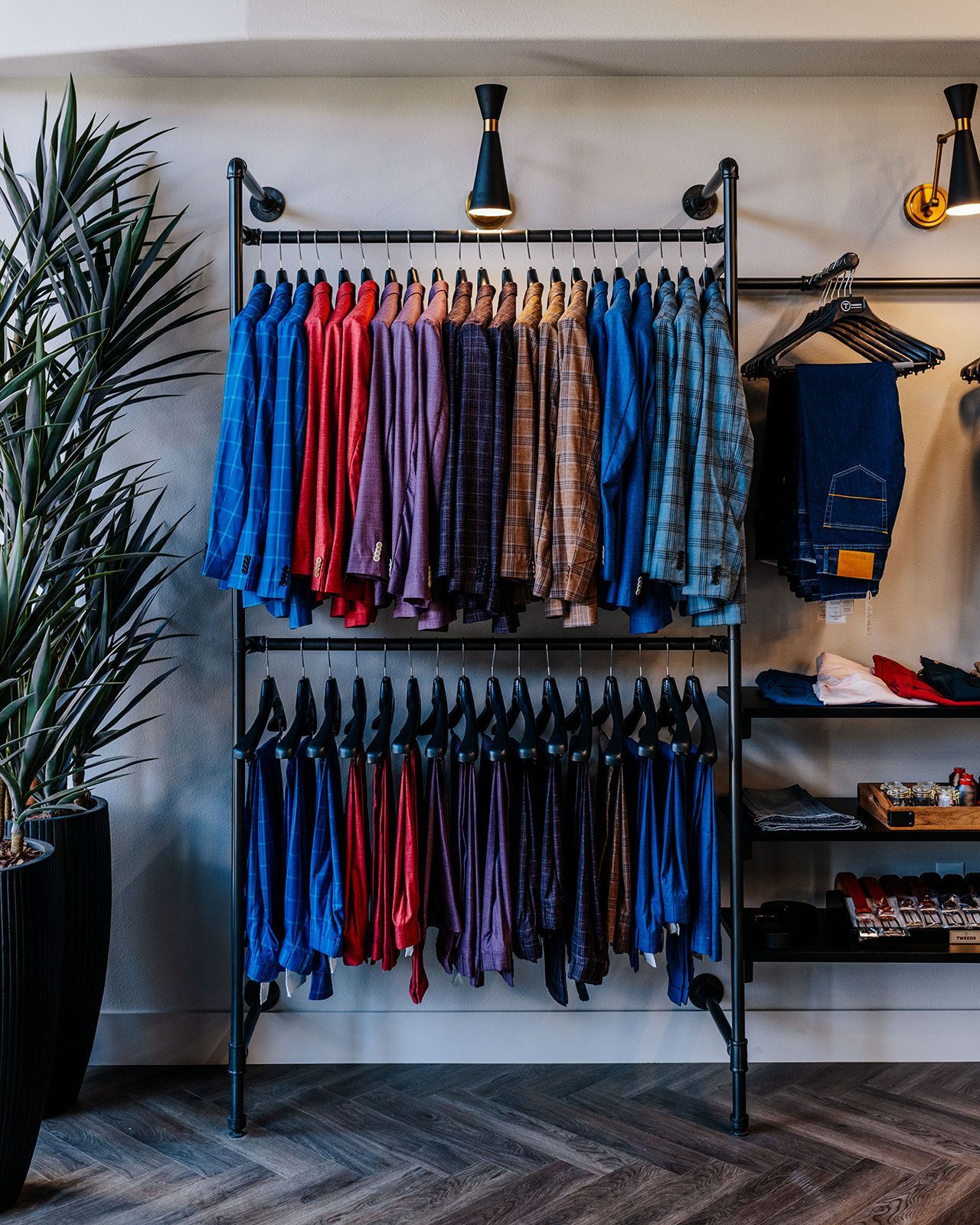Clothing rack in a store, displaying colorful shirts and pants, with a plant to the left.