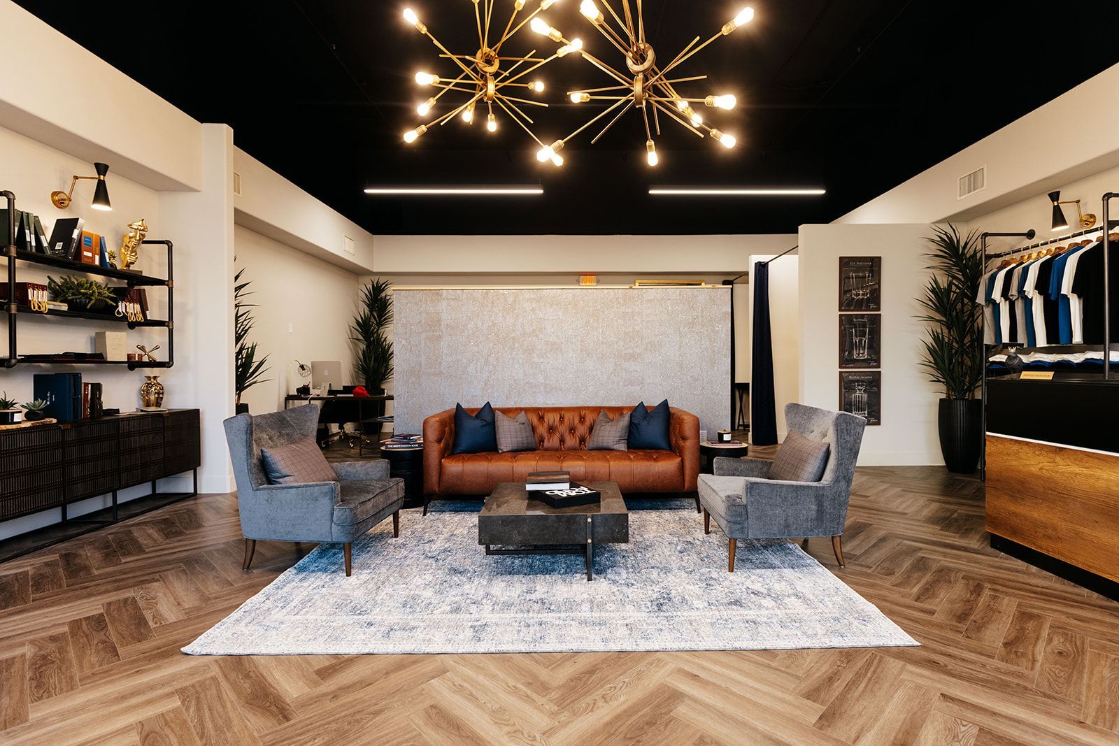 A modern clothing store interior with brown leather sofa, armchairs, black shelving and ceiling, and a sparkling chandelier.
