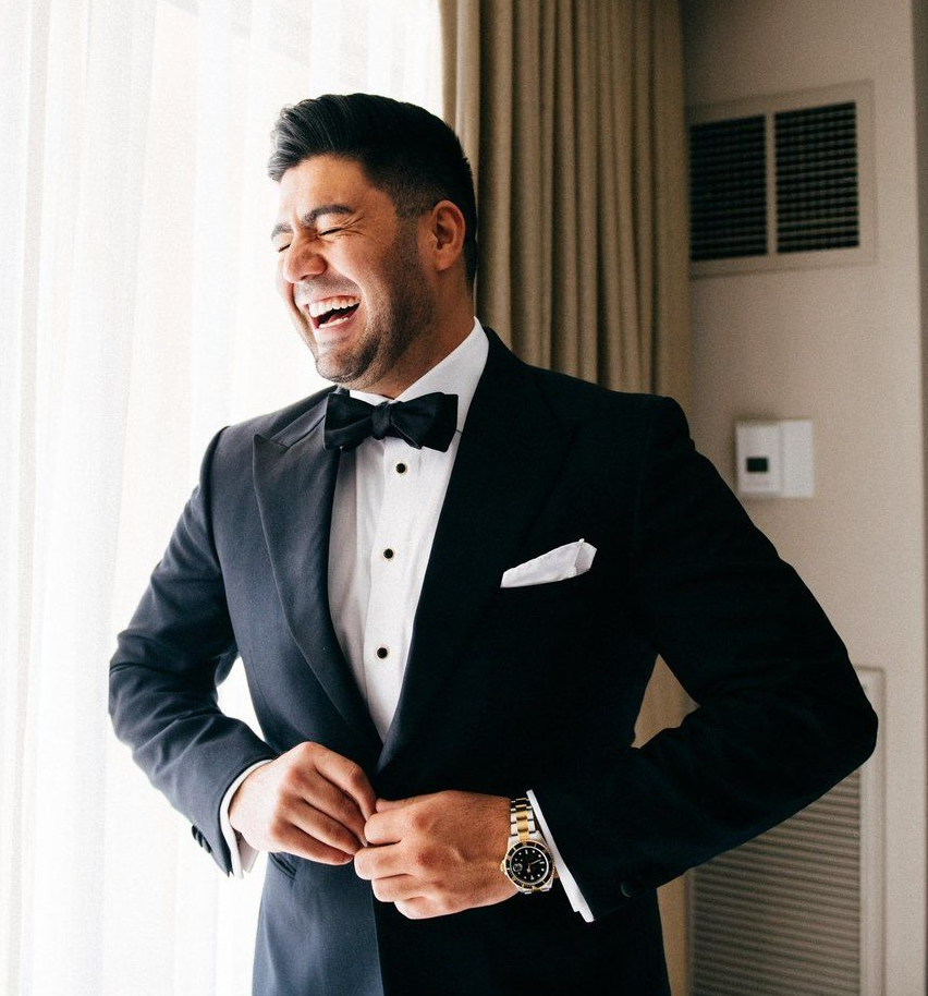 Man in a tuxedo laughs, adjusting his jacket near a window. Gold watch visible.