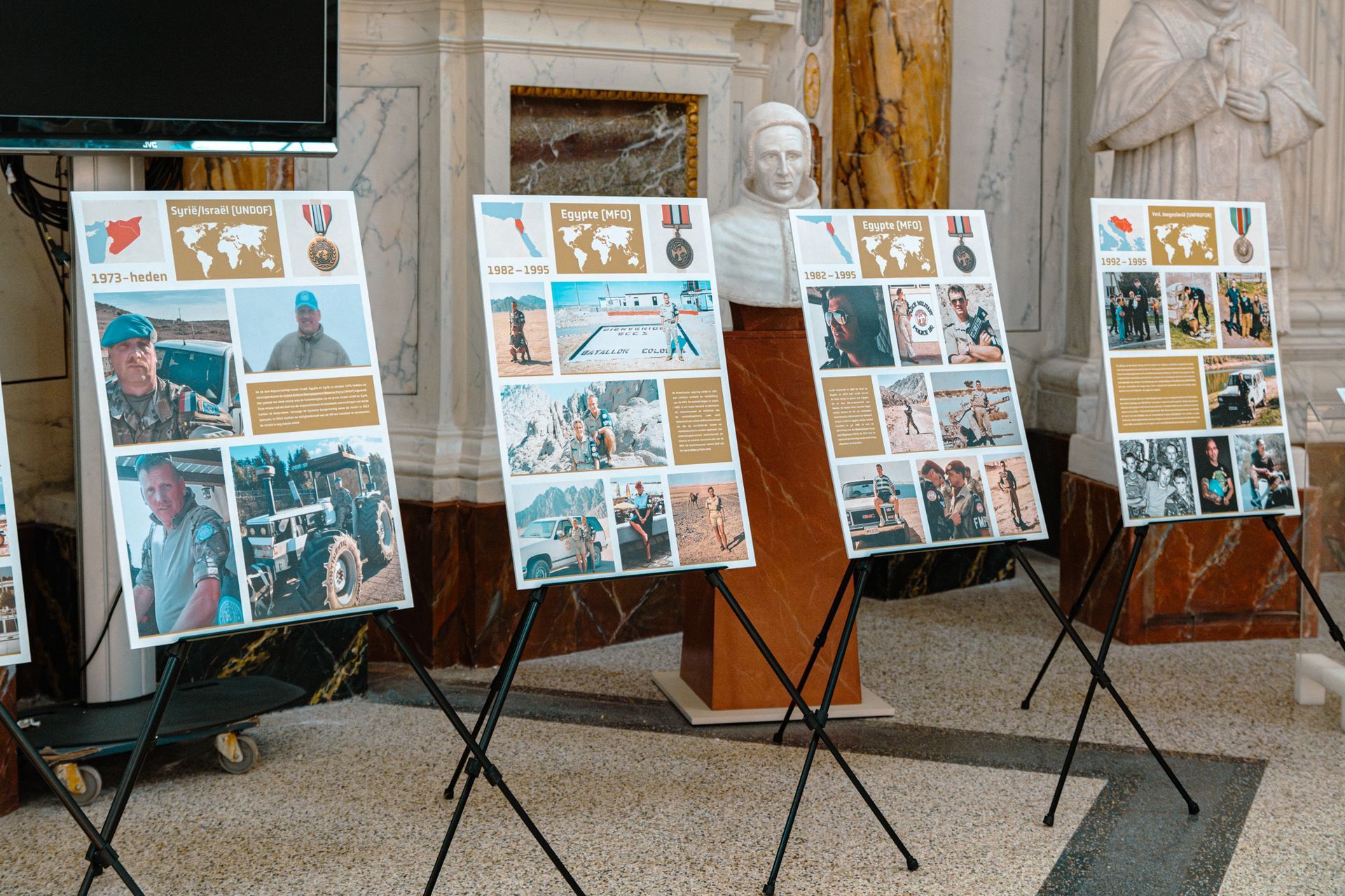In een hal staan ​​vier informatieve posters op schildersezels, elk met foto's, kaarten en symbolen over militaire missies.