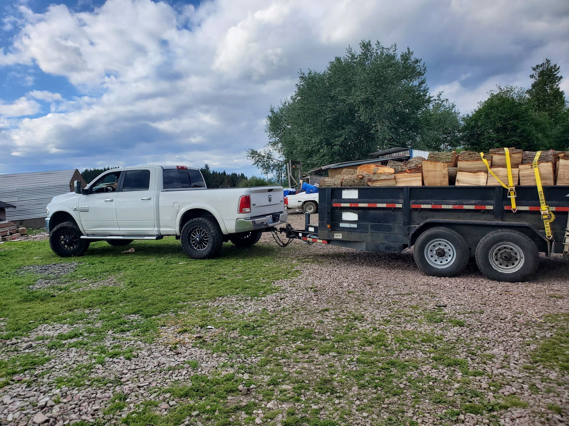 White pickup truck towing a trailer loaded with firewood on a grassy field under a cloudy sky.