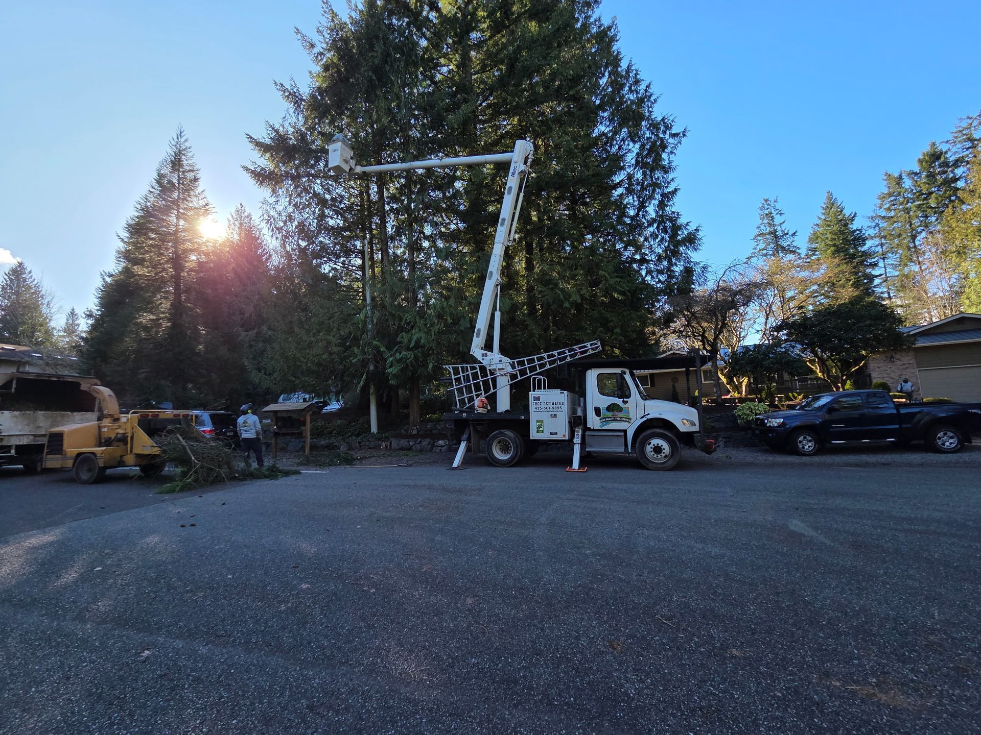 Bucket truck with raised boom in a gravel lot among trees and parked cars at sunset