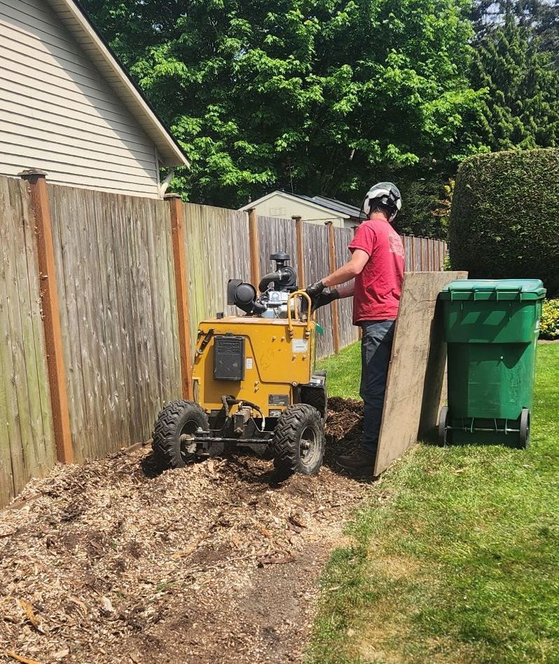 Person using a yellow machine to remove a wooden fence panel in a backyard beside green trash bins.