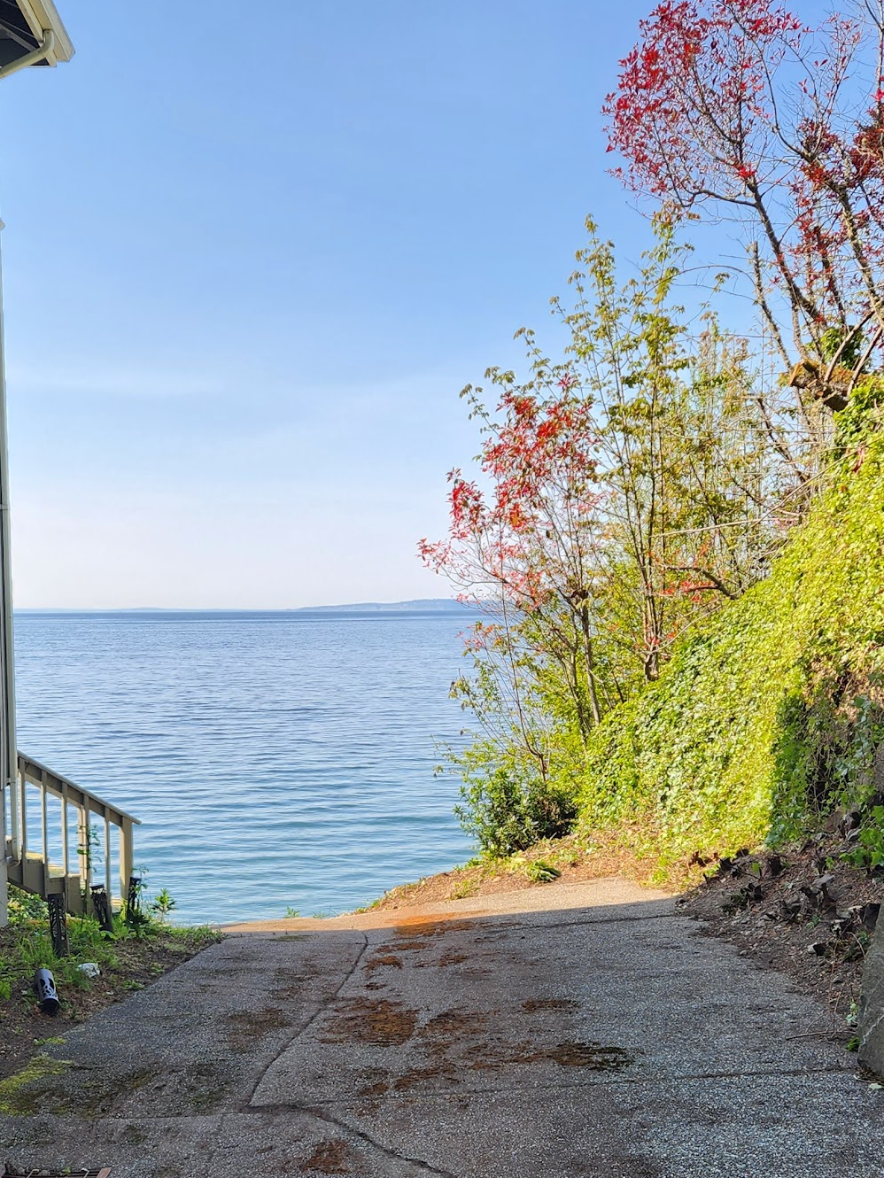 Gravel path slopes down to a calm body of water. Trees with red and green leaves frame the water. Clear blue sky.
