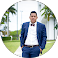 Man in a navy suit standing outdoors beside a palm tree and modern building.