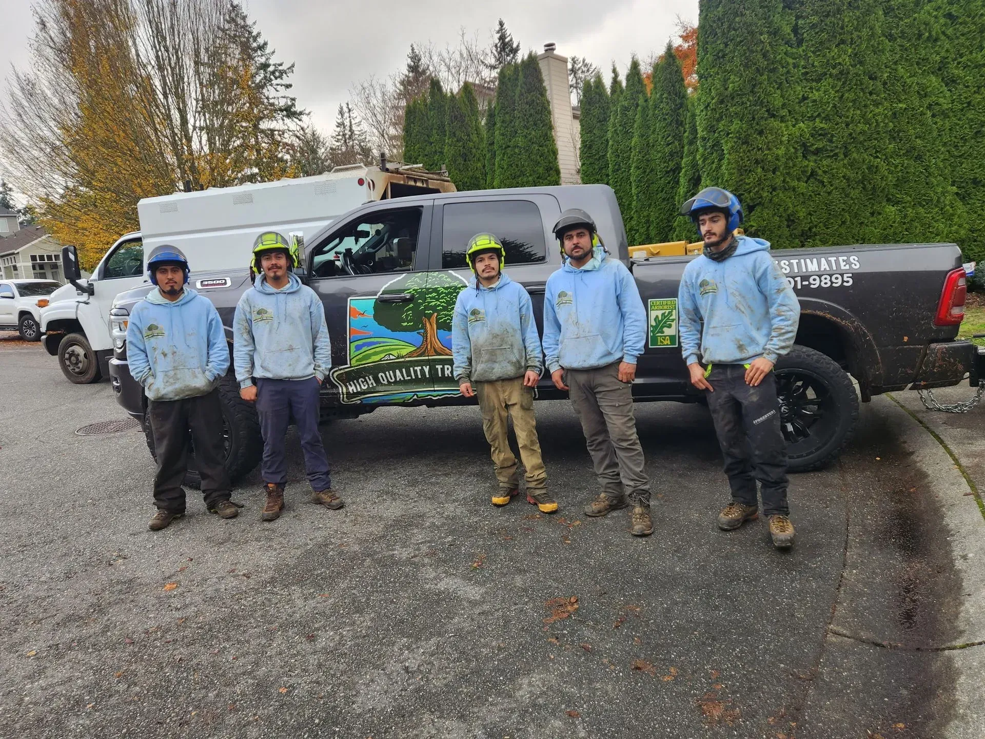 Five people in blue jackets stand beside a wrapped truck in a gravel parking area, with trees in the background.