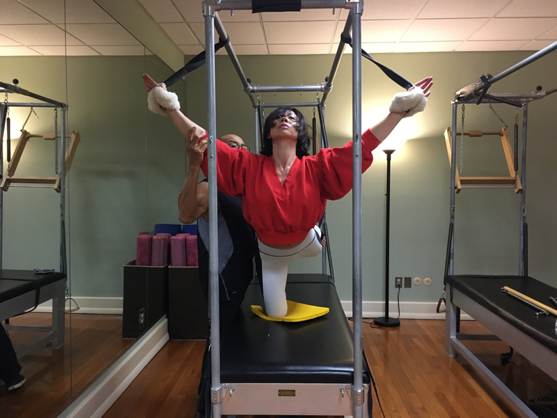 Woman doing Pilates exercise with assistance from a person, on a reformer machine. Room has mirrors and equipment.